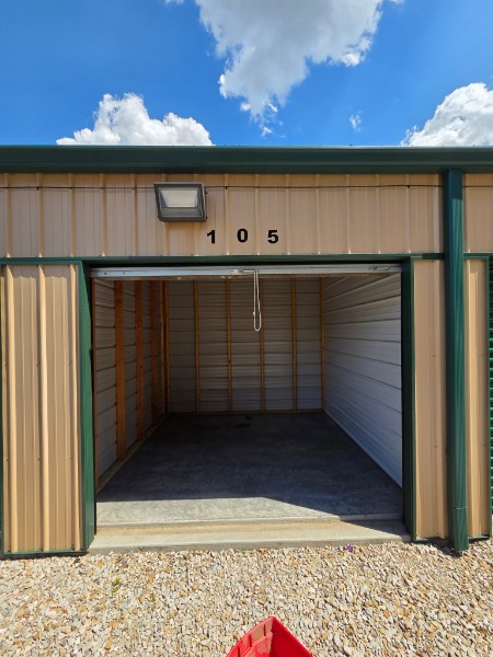 Exterior view of storage unit 105 with open roll-up door, security light above, and blue sky in the background